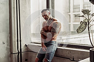 Fashion portrait of naked male model with tattoo and a black beard standing in hot pose on near the window. Loft room interio