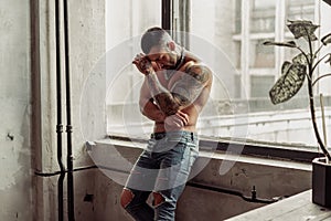 Fashion portrait of naked male model with tattoo and a black beard standing in hot pose on near the window. Loft room interio