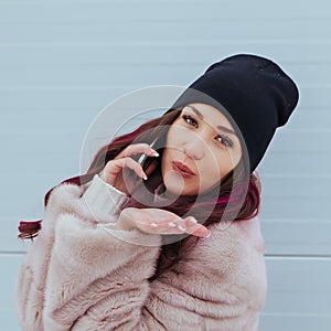 Fashion portrait of pretty smiling hipster woman in mink coat and smartphone against the colorful grey wall. Talking