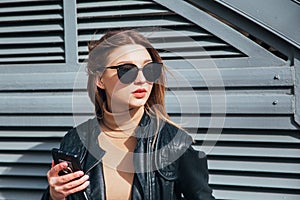 Fashion portrait pretty woman in black rock style in sunglasses using mobile phone over gray background in city