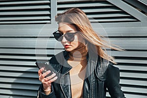 Fashion portrait pretty woman in black rock style in sunglasses using mobile phone over gray background in city