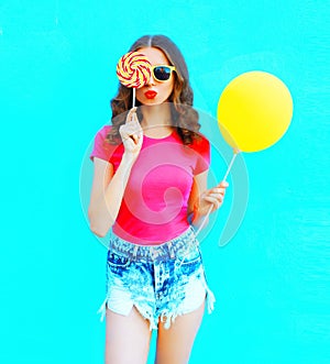 Fashion portrait pretty young woman wearing pink t-shirt, denim shorts with yellow air balloon, lollipop candy over colorful blue