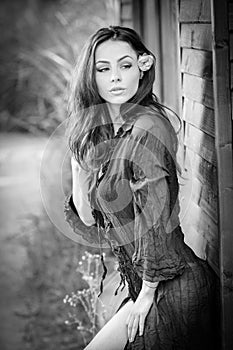 Fashion portrait of brunette in black blouse leaning on wooden cabin wall. Sensual attractive woman with a flower in hair