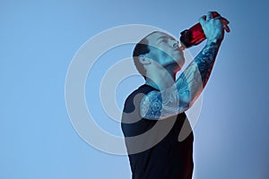 Fashion portrait of sport fit attractive man drinking water from bottle, tattooed hands, hipster look. Color flash studio light