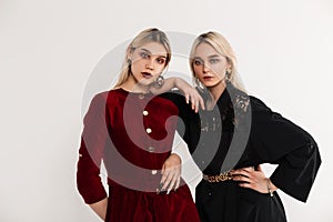 Fashion portrait young attractive blondes sisters women in red and black fashionable dresses near white vintage wall in studio.