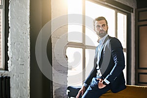 Fashion portrait. Young bearded successful businessman in stylish suit is standing at office and thinking about