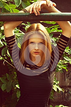 Fashion portrait of young beautiful red-haired girl wearing trendy black casual dress and posing over green tree with hands up.