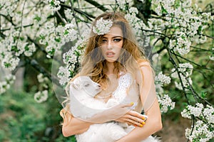 Fashion portrait of young beautiful woman holding a small lamb in spring blooming garden.