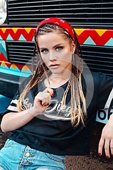 Fashion portrait of young hipster woman with bandana and long braids sitting on city street