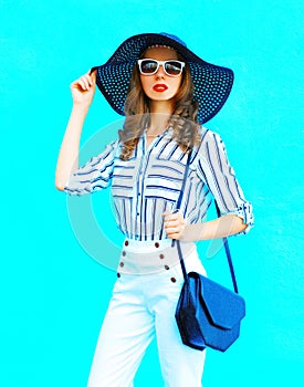 Fashion portrait young woman wearing a straw hat, white pants and handbag clutch over colorful blue background posing in city