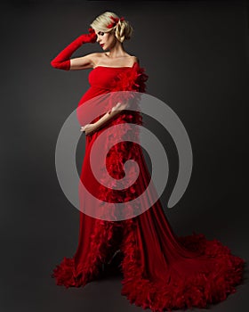 Fashion Pregnant Woman in Luxury Red Dress with Feather. Elegant Mother in evening Gown looking at Belly Side view. Pregnancy