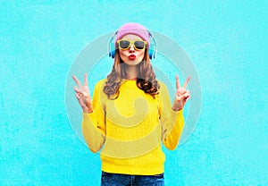 Fashion pretty cool girl in headphones listening to music wearing colorful pink hat yellow sunglasses and sweater over blue