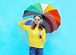 Fashion pretty cool girl listens to music in headphones with colorful umbrella in autumn day over colorful blue background