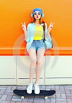 Fashion pretty cool girl is listens to music with a skateboard over colorful orange