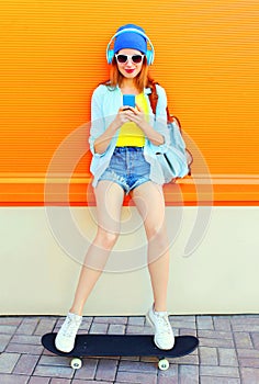 Fashion pretty cool girl is listens to music and using a smartphone sits on a skateboard over colorful orange