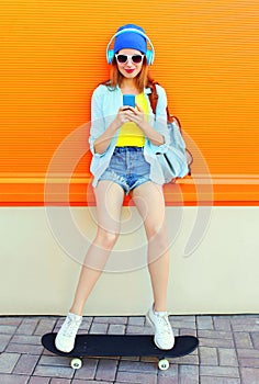 Fashion pretty cool girl listens to music using smartphone on skateboard over colorful orange