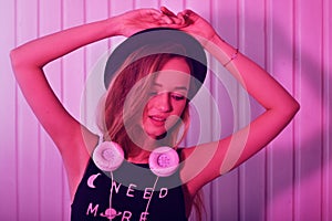 Fashion pretty cool woman in hat and headphones listening to music over pink neon background. Beautiful young teenage girl in hat