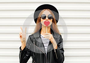 Fashion pretty cool young girl with red lollipop heart wearing black hat leather jacket over white urban