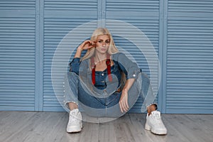 Fashion pretty stylish woman in fashionable denim clothes with white sneakers sits near the blue wooden wall.