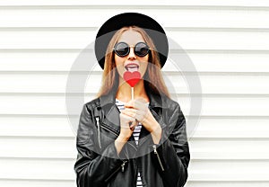 Fashion pretty sweet young woman enjoying a taste lollipop heart wearing black hat leather jacket over white