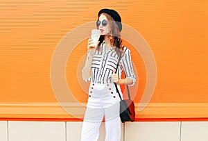 Fashion pretty woman with coffee cup wearing a black hat white pants handbag clutch over colorful orange