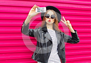 Fashion pretty woman makes self portrait on smartphone in black rock style over city pink