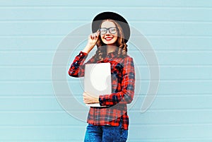 Fashion pretty young smiling woman holding laptop computer or tablet pc in city, wearing a black hat, red checkered shirt