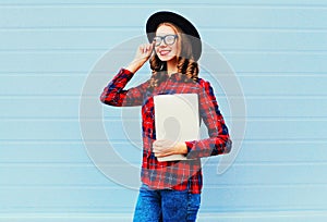 Fashion pretty young smiling woman holding laptop computer or tablet pc in city, wearing black hat, red checkered shirt over blue