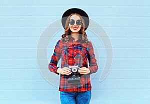 Fashion pretty young smiling woman holds retro camera wearing black hat red checkered shirt over blue background