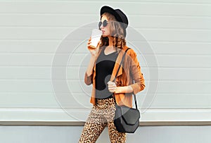 Fashion pretty young woman drinks coffee of cup wearing a retro elegant hat, sunglasses, brown jacket and black handbag