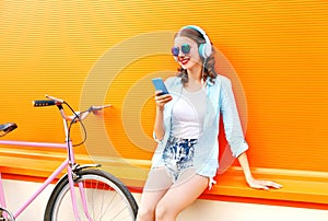 Fashion pretty young woman listens to music using smartphone near urban retro bicycle over colorful orange