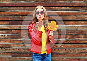 Fashion pretty young woman sends air sweet kiss with yellow maple leafs in autumn day over wooden background wearing red leather