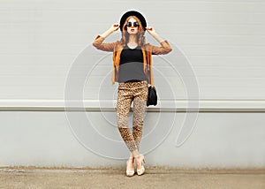 Fashion pretty young woman wearing a retro elegant hat, sunglasses, brown jacket and black handbag posing in city over backgr