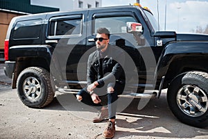 Fashion rich beard Arab man wear on black jeans jacket and sunglasses posed against big black suv car. Stylish, succesful and