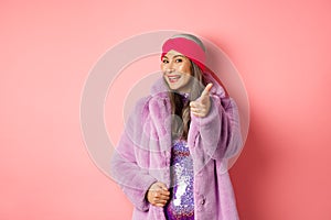 Fashion and shopping concept. Happy asian mature woman in stylish fake-fur coat pointing finger at camera, we need you