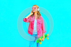 Fashion smiling woman holds skateboard listens to music in wireless headphones in pink denim jacket