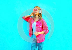 Fashion smiling woman listens to music in wireless headphones in pink denim jacket on blue