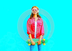 Fashion smiling woman with a skateboard listens to music in wireless headphones in pink denim jacket