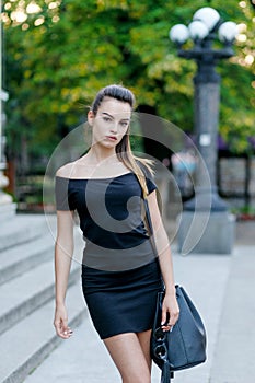 Fashion style portrait of young beautiful calm female model posing at city street in black slim dress and bag.