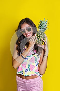 Fashion summer portrait smiling indian girl in sunglasses and pineapple over yellow background