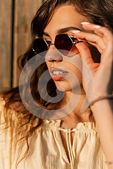 Fashion summer portrait of stylish pretty woman with curly hair put on a sunglasses near wooden wall at sunset