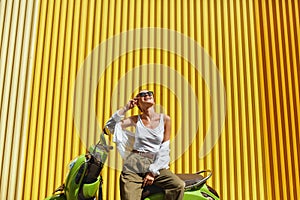 Fashion. Urban Girl On Moped Portrait. Stylish Sexy Woman In Casual Clothes And Sunglasses Against Yellow Metal Fence