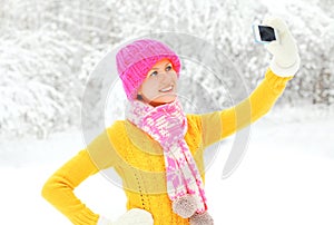 Fashion winter happy smiling young woman taking picture self portrait on smartphone over snowy trees wearing colorful knitted hat