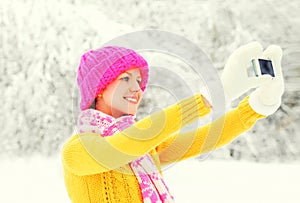 Fashion winter happy smiling young woman taking picture self portrait on smartphone over snowy trees wearing colorful knitted hat