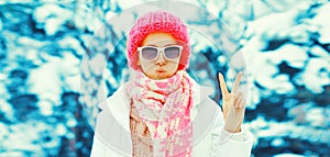 Fashion winter portrait of stylish young woman wearing sunglasses, colorful knitted hat and scarf in the park on snowy background