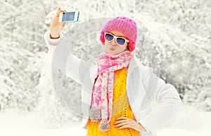 Fashion winter woman taking photo self portrait on smartphone over snowy trees wearing a colorful knitted hat scarf
