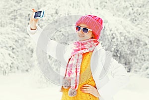 Fashion winter woman taking picture self portrait on smartphone over snowy trees wearing a colorful knitted hat scarf