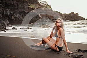 Fashion woman in black swimwear relaxing on tropical beach, enjoying sunbath near the rocky sea shore