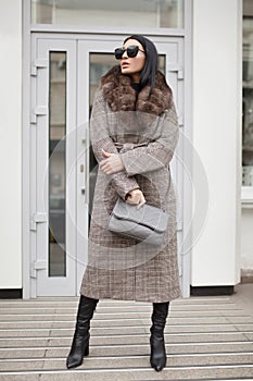 Fashion woman in the city stands in front of a white door. She is wearing a coat. sunglasses and bag