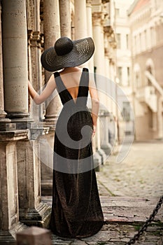 Fashion Woman Model in Hat Back Side View on Old City Street. Elegant Traveler Girl in Black Dress Looking at Antique Architecture